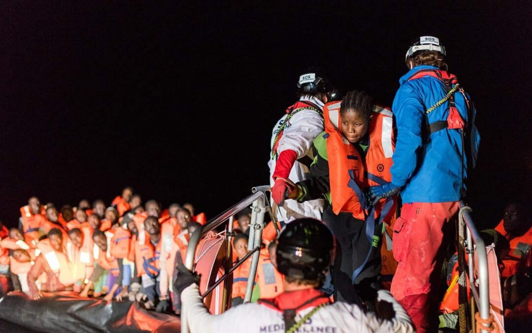 Photo de nuit, sur l'eau. Au premier plan: une femme rescapée est aidée par des marins-sauveteurs à monter sur le canot de sauvetage. Au second plan: un bateau pneumatique surchargé de rescapés en gilets de sauvetage.